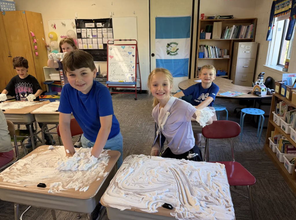 Several second-grade students smile and pose in their classroom while using shaving cream spread across their desks to practice writing and drawing. Some students hold shaving cream in their hands while others create designs on their desks.