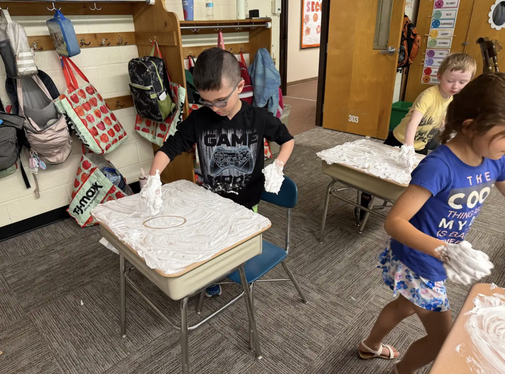 A student wearing glasses carefully traces shapes and letters in a layer of shaving cream spread across a desk while classmates participate in the activity around the classroom.