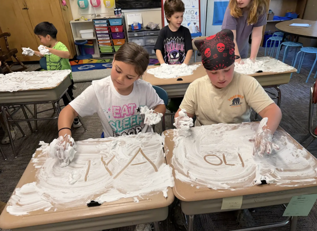 Two second-grade students concentrate on writing letters and words in shaving cream spread across their desks. Other classmates can be seen working on the same activity in the background.