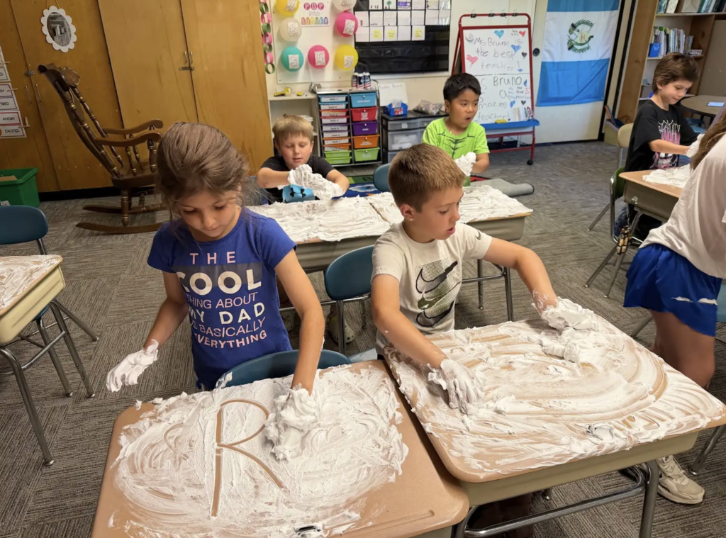Students sit at their desks covered in shaving cream, practicing letter formation and drawing shapes with their fingers while classmates work nearby.