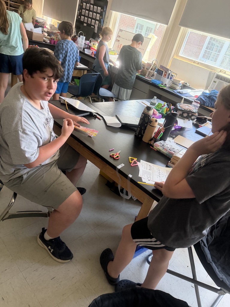 Students gather around a science lab table, measuring, taping, and constructing straw-based geometric designs during a STEM challenge.