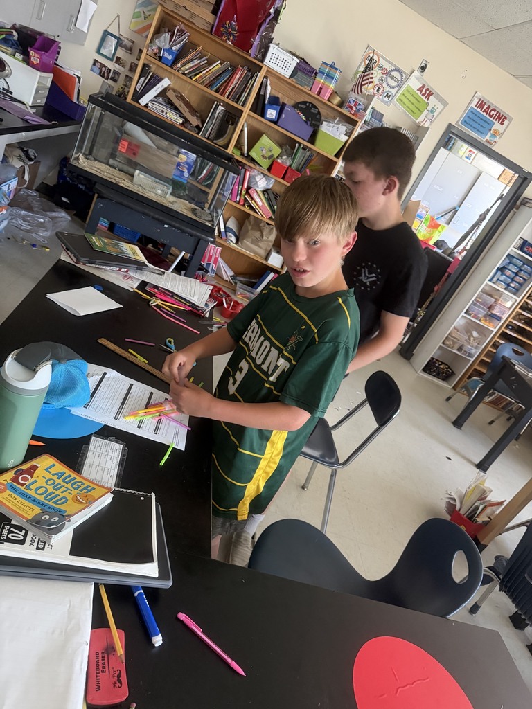 Two students stand at a classroom table arranging colorful straws and tape while planning the next steps of their engineering project.