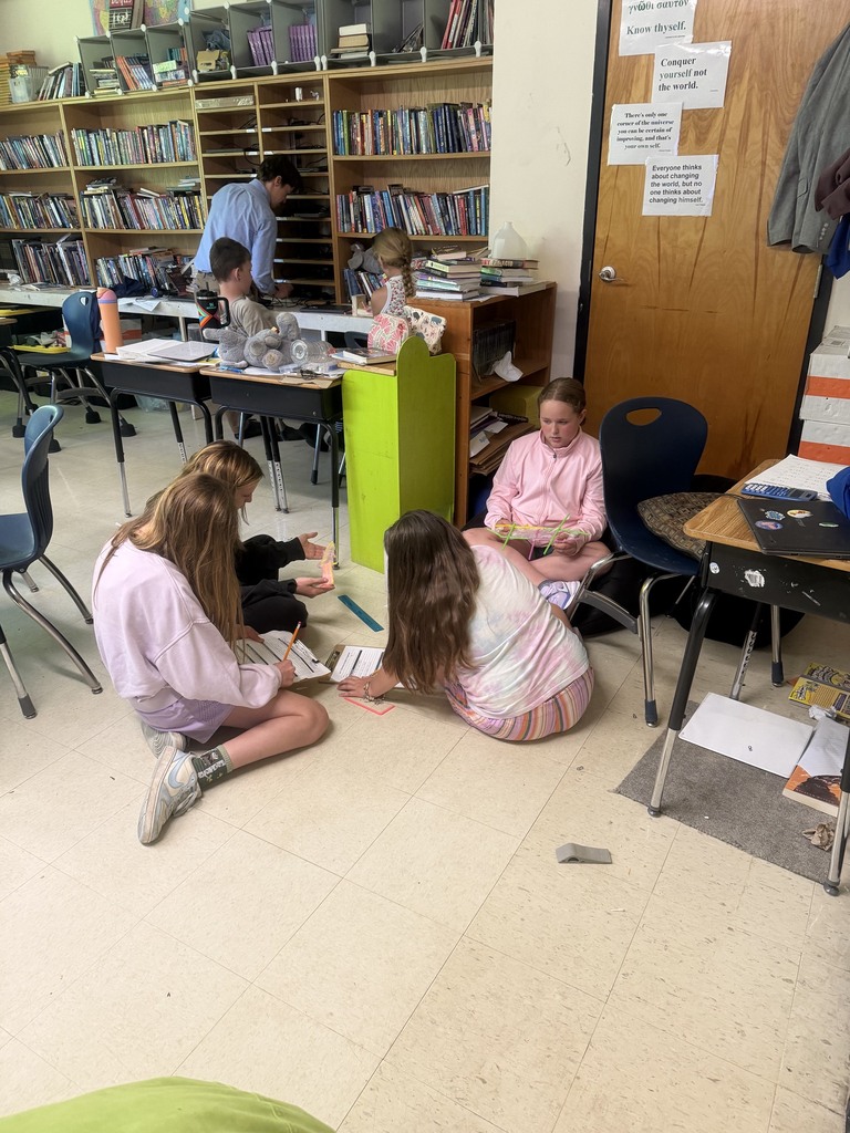 A small group of students sits on the classroom floor discussing and planning their straw structure design while completing a STEM engineering challenge.