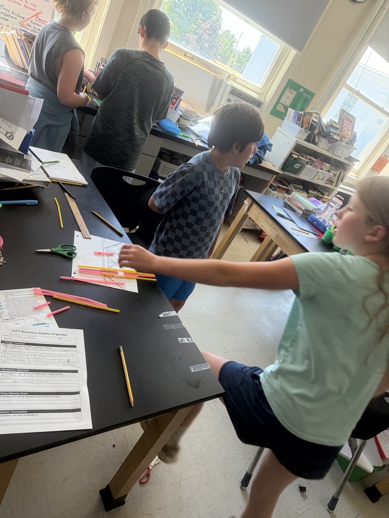 Several students collaborate around a classroom table covered with rulers, scissors, and colorful straws as they test and refine their structure design.