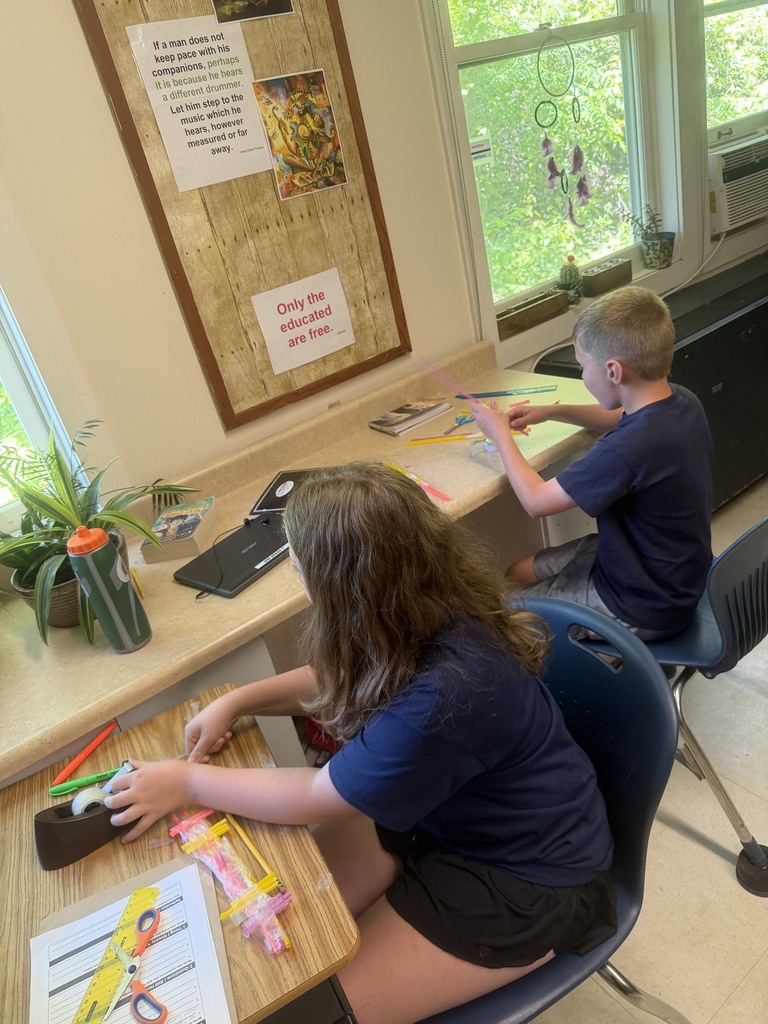 Two students sit by a classroom window assembling a structure from colorful straws, rulers, and tape during a collaborative engineering activity.
