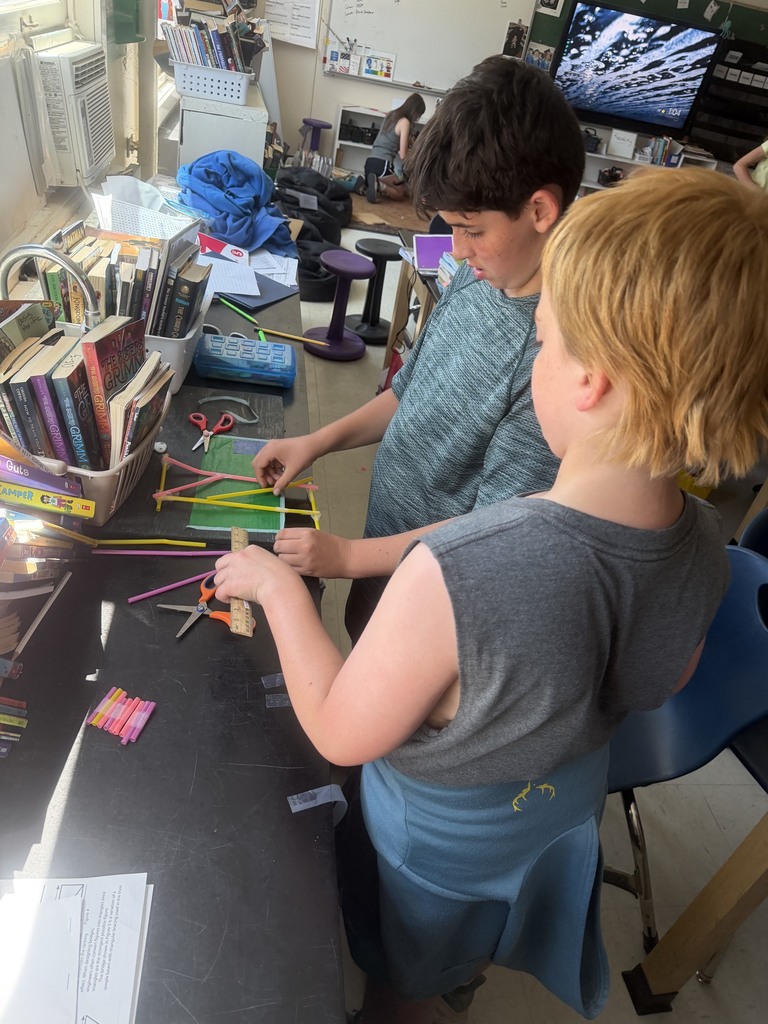 Two students examine a partially completed straw framework on a classroom table, measuring and adjusting pieces as part of a STEM building activity.