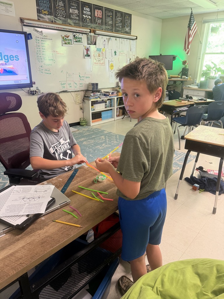 Two students work together at a classroom table, using colorful drinking straws and tape to build a geometric structure as part of a hands-on STEM activity.