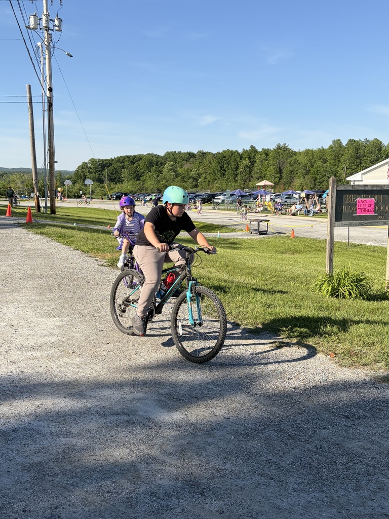 A wide shot of the school's "Bike and Groove" event. Dozens of students are riding their bicycles along a paved path winding through the school campus, surrounded by green grass [and trees/school buildings].