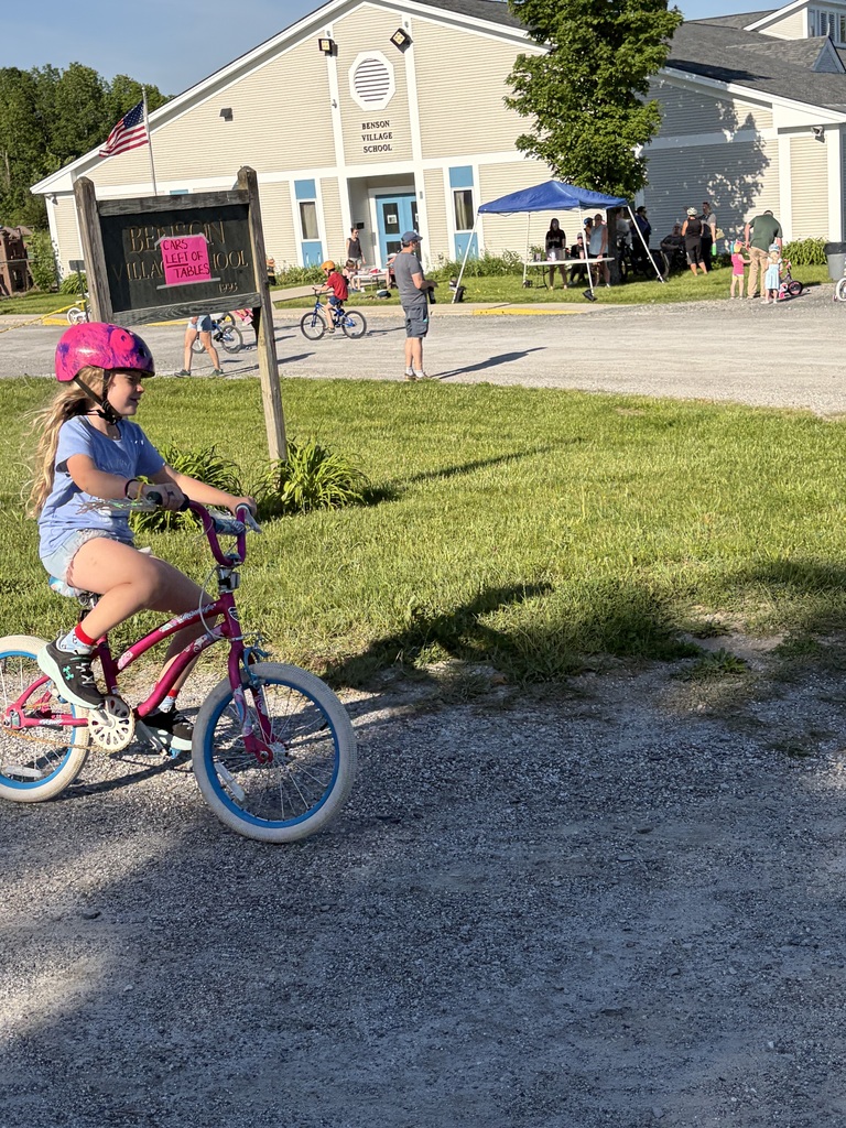 A wide shot of the school's "Bike and Groove" event. Dozens of students are riding their bicycles along a paved path winding through the school campus, surrounded by green grass [and trees/school buildings].