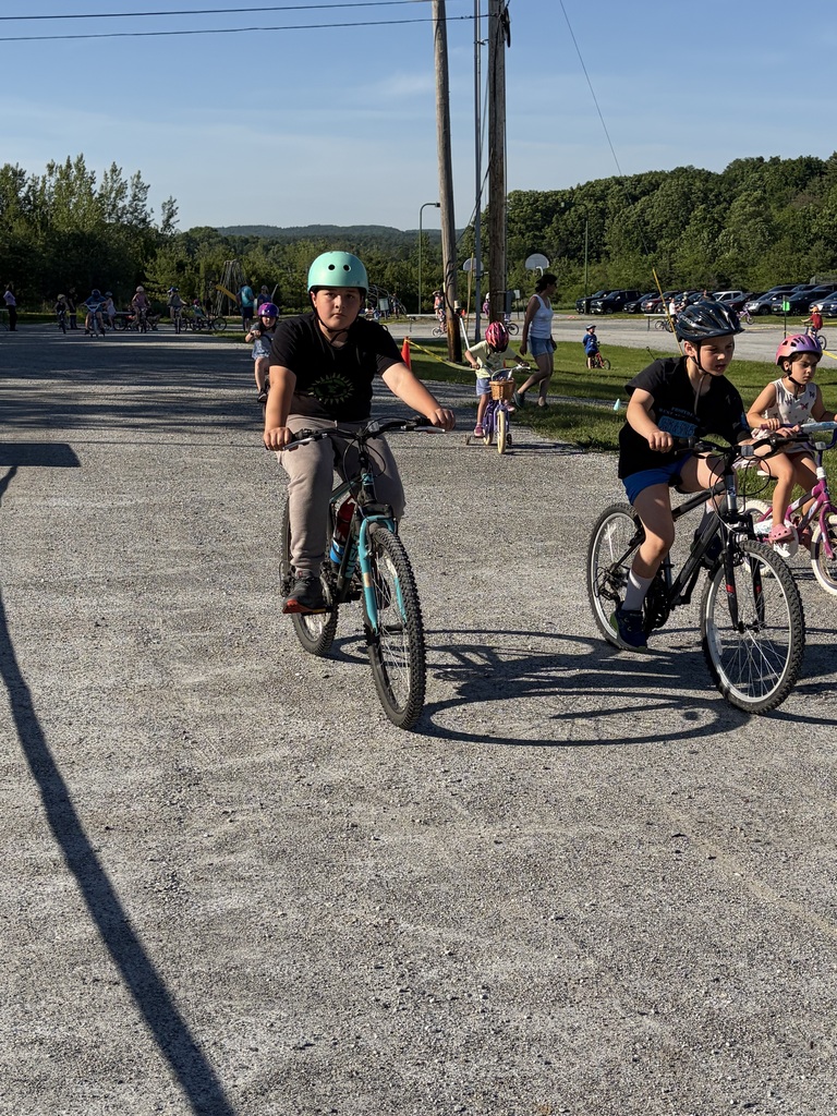 A wide shot of the school's "Bike and Groove" event. Dozens of students are riding their bicycles along a paved path winding through the school campus, surrounded by green grass [and trees/school buildings].