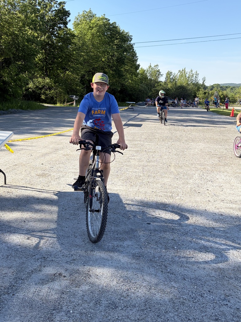 A wide shot of the school's "Bike and Groove" event. Dozens of students are riding their bicycles along a paved path winding through the school campus, surrounded by green grass [and trees/school buildings].
