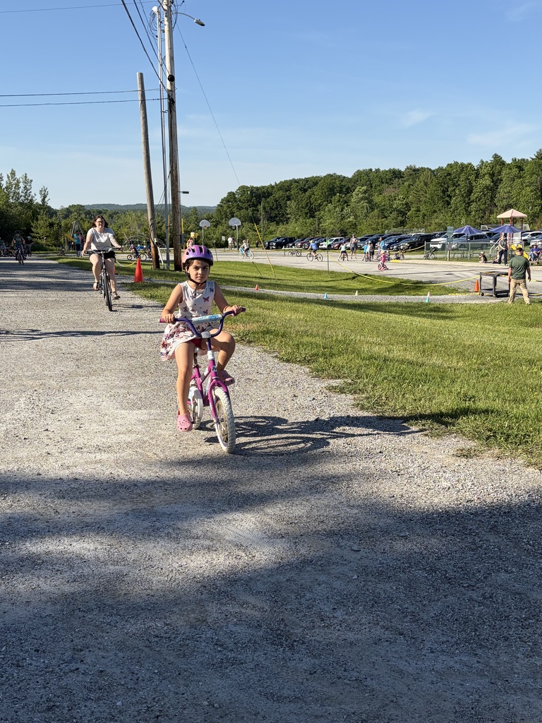 A wide shot of the school's "Bike and Groove" event. Dozens of students are riding their bicycles along a paved path winding through the school campus, surrounded by green grass [and trees/school buildings].