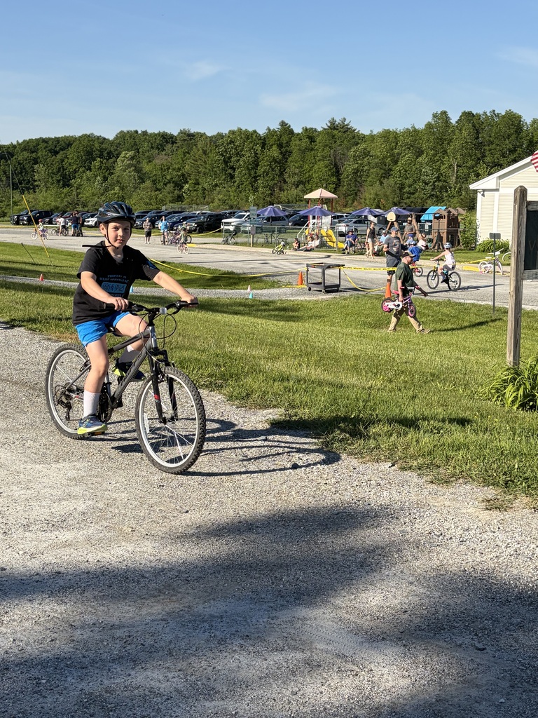 A wide shot of the school's "Bike and Groove" event. Dozens of students are riding their bicycles along a paved path winding through the school campus, surrounded by green grass [and trees/school buildings].