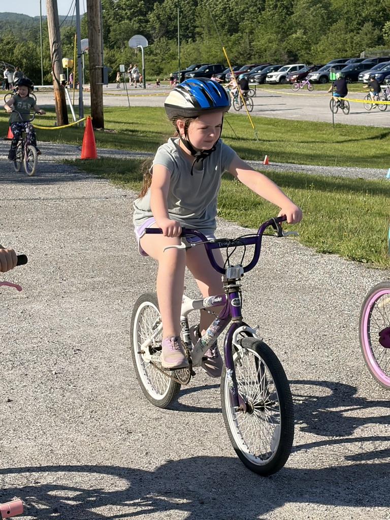 A wide shot of the school's "Bike and Groove" event. Dozens of students are riding their bicycles along a paved path winding through the school campus, surrounded by green grass [and trees/school buildings].