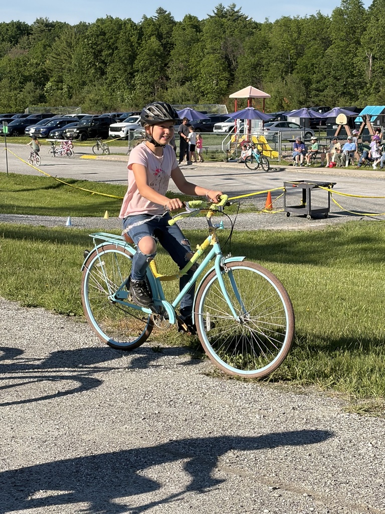 A young girl with a black helmet and a pink shirt smiles while riding a light blue cruiser bicycle on a gravel path. The bike is decorated with yellow streamers on the handlebars and frame. In the background, there is a grassy field with a playground, parked cars, and other people.