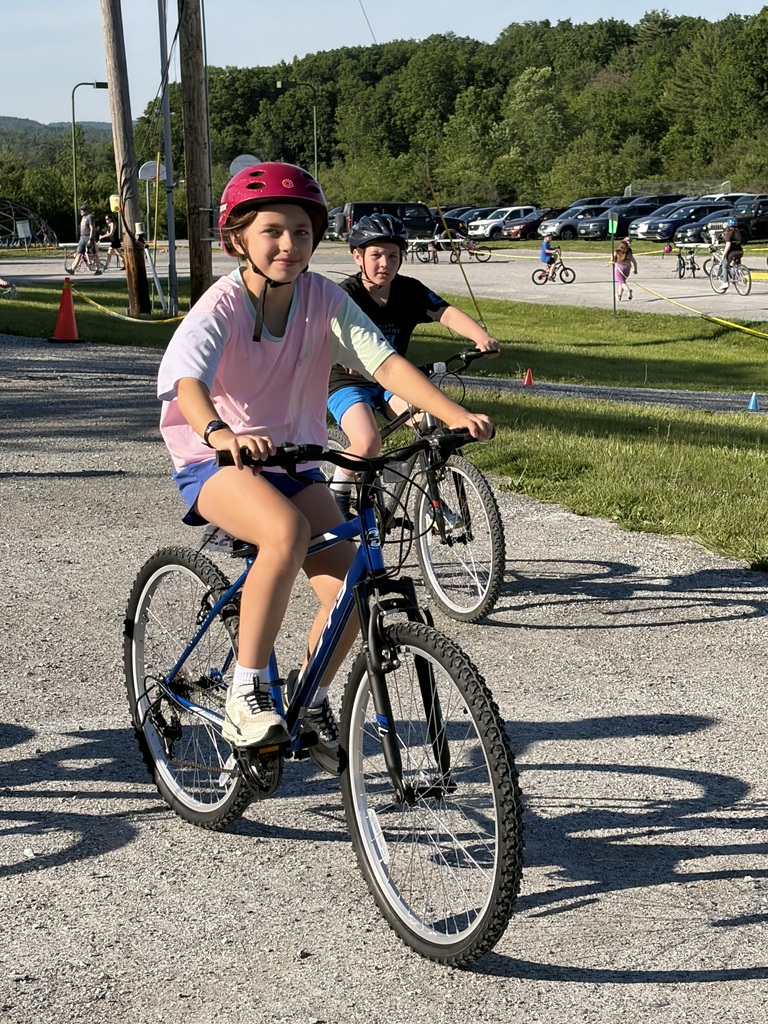 A young child wearing a bright pink helmet and a light yellow t-shirt rides a purple bicycle with training wheels and a wicker front basket. The child is riding on a gravel driveway in front of a building with a sign that reads "Benson Village School."