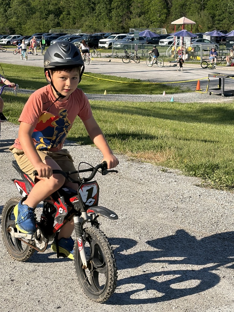 A young boy wearing a large black helmet and an orange graphic t-shirt rides a black and red youth bicycle designed to look like a motocross dirt bike. He is riding on a gravel path with a sunny playground and parked cars in the background. IMG_0586.jpg Alt Text: A smiling girl wearing a pink helmet and a pink and white color-blocked t-shirt rides a blue mountain bike on a gravel path. Another boy on a bicycle follows closely behind her, with a parking lot and tree line under a bright sky in the background.