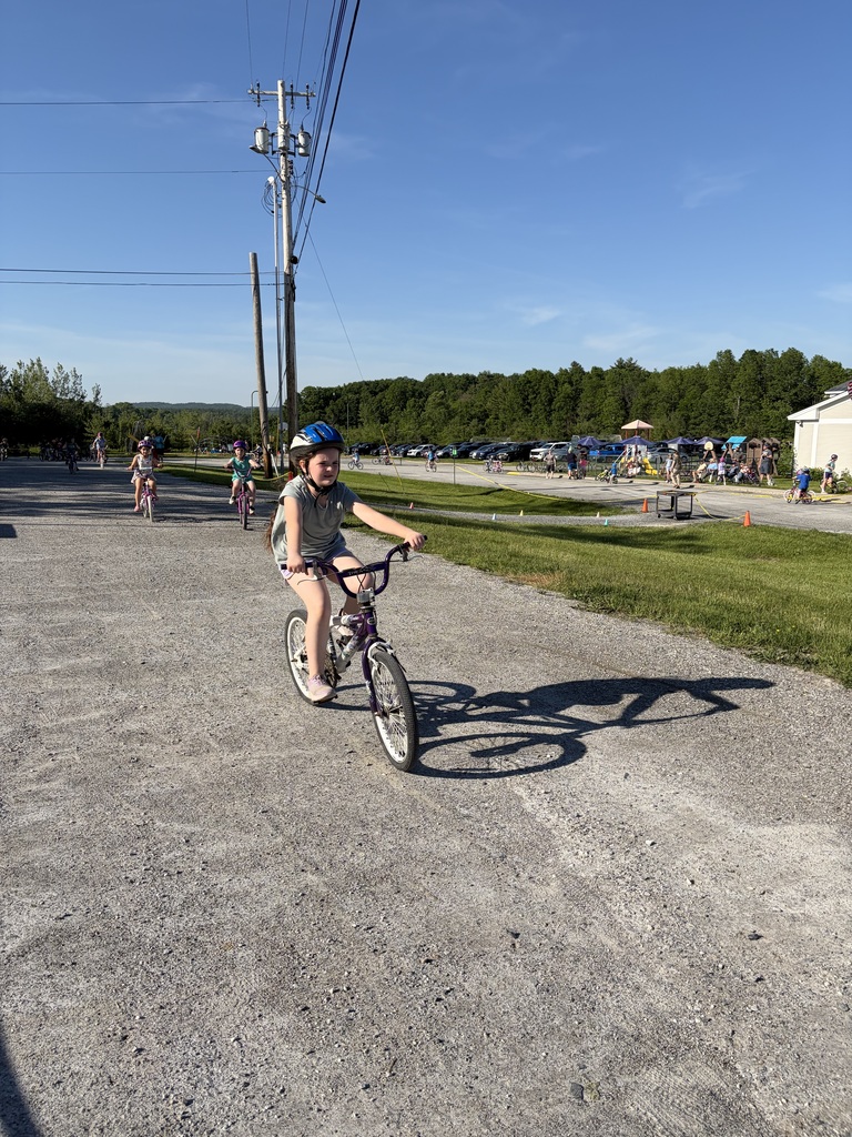 A girl wearing a blue helmet and a grey t-shirt rides a purple mountain bike down a wide gravel path, casting a long shadow in front of her. Other children on bicycles follow behind her under a clear blue sky, with a playground and parking lot visible in the distance.