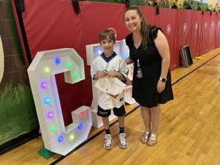 Student holds a certificate and medal while standing with a staff member near lighted display letters.