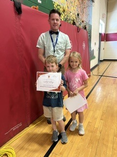 Student receives a certificate while standing with two classmates and a staff member in the school gym.