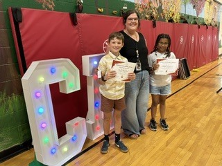 Two students display their certificates while posing with a staff member beside illuminated letter decorations.