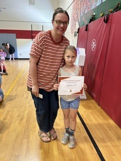 Student smiles while holding a certificate next to a staff member in the gym.