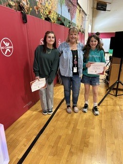 Two students proudly display certificates while standing with a staff member in the school gym.