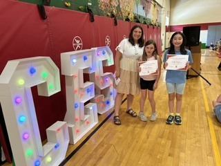 Two students display certificates while standing with a staff member beside illuminated “CES” letters.
