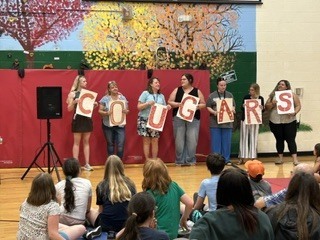 Staff members hold large letters spelling “COUGARS” in front of seated students during a school assembly.