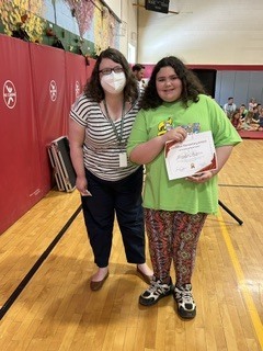 A student smiles while holding a certificate and posing with a staff member during a school awards ceremony.