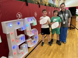 Two students hold certificates while posing with a staff member beside illuminated “CES” letters during a school recognition ceremony.