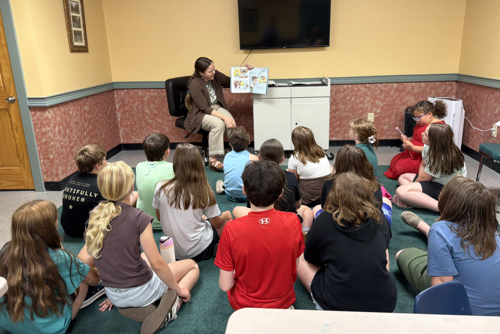 A librarian reads aloud from a picture book to a group of fourth-grade students seated on the floor at the Fair Haven Public Library.