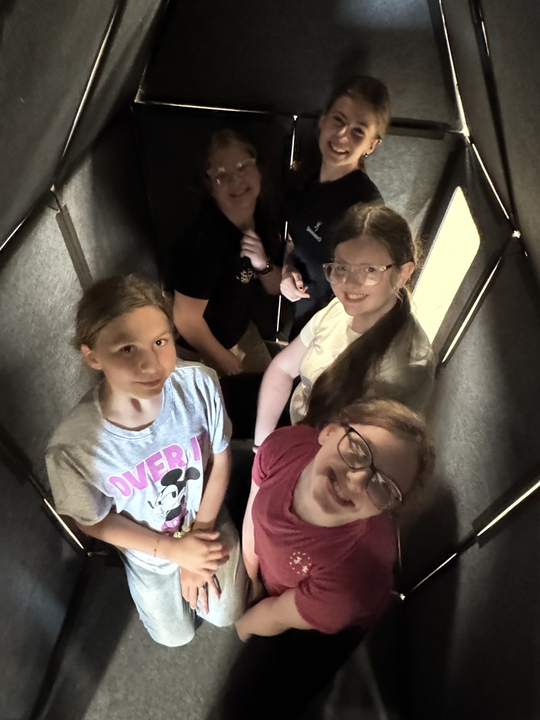 Five children standing inside a small gray panel fort or enclosure, looking up and smiling at the camera. A boy sits inside a gray geometric play structure on a classroom floor, looking up at the camera. A smiling boy with glasses sits inside a larger gray panel structure while other children assemble pieces around him. Several children build with large gray modular panels in a gym or classroom space with blue wall mats in the background. Three children work together to attach triangular and rectangular gray panels into a tall structure while others move nearby.