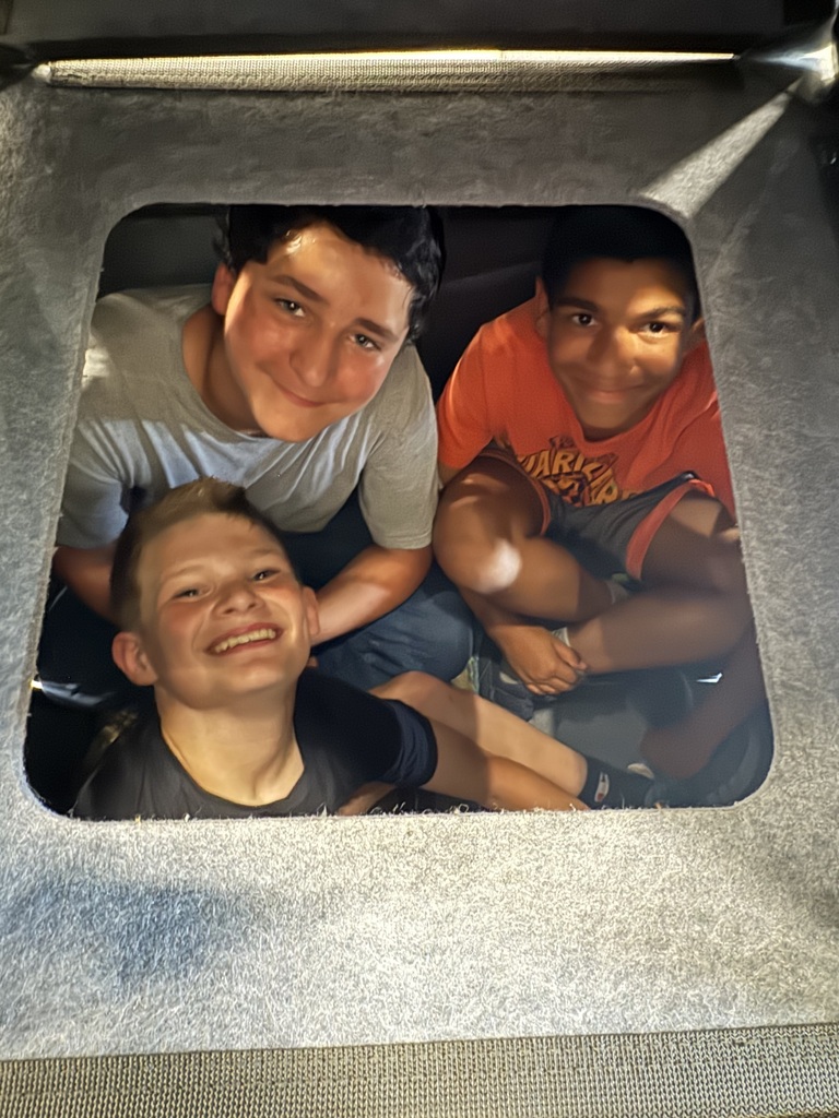 A close-up shot looking through a square window cutout of a grey modular structure, framing three smiling boys crowded together inside. The boy on the left wears a grey shirt, the boy in the center foreground wears a black shirt and smiles widely with tilted head, and the boy on the right wears a bright orange t-shirt. The interior of the fort is dimly lit, creating a cozy clubhouse atmosphere.