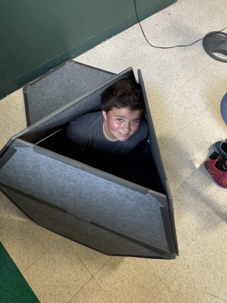 A boy sits inside a gray geometric play structure on a classroom floor, looking up at the camera.