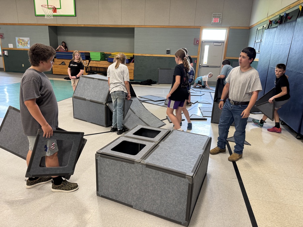 A wide shot inside a school gymnasium where a group of about nine middle-school-aged children are working together to assemble large, modular building structures. The building pieces are made of thick, grey felt-like panels with dark grey borders. In the foreground, a large rectangular base has two square window cutouts on top. To the left, a boy carries a panel with a window cutout. In the background, other students attach panels to form fort-like structures, while extra triangular and rectangular pieces lie scattered across the gym floor.