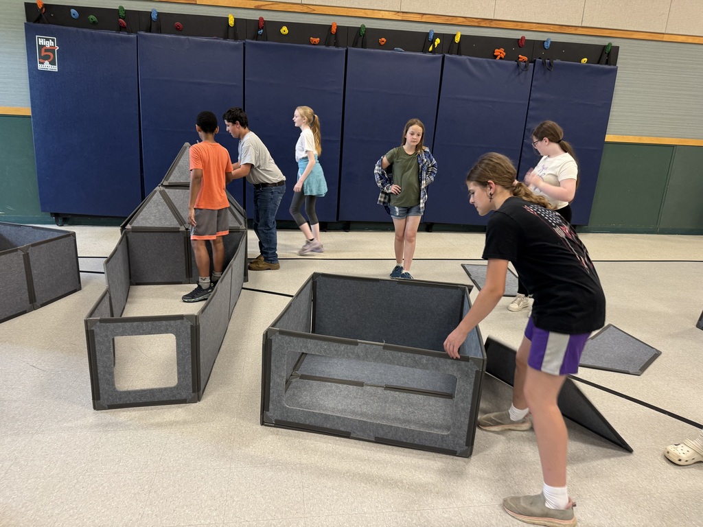 Several children build with large gray modular panels in a gym or classroom space with blue wall mats in the background. Three children work together to attach triangular and rectangular gray panels into a tall structure while others move nearby.