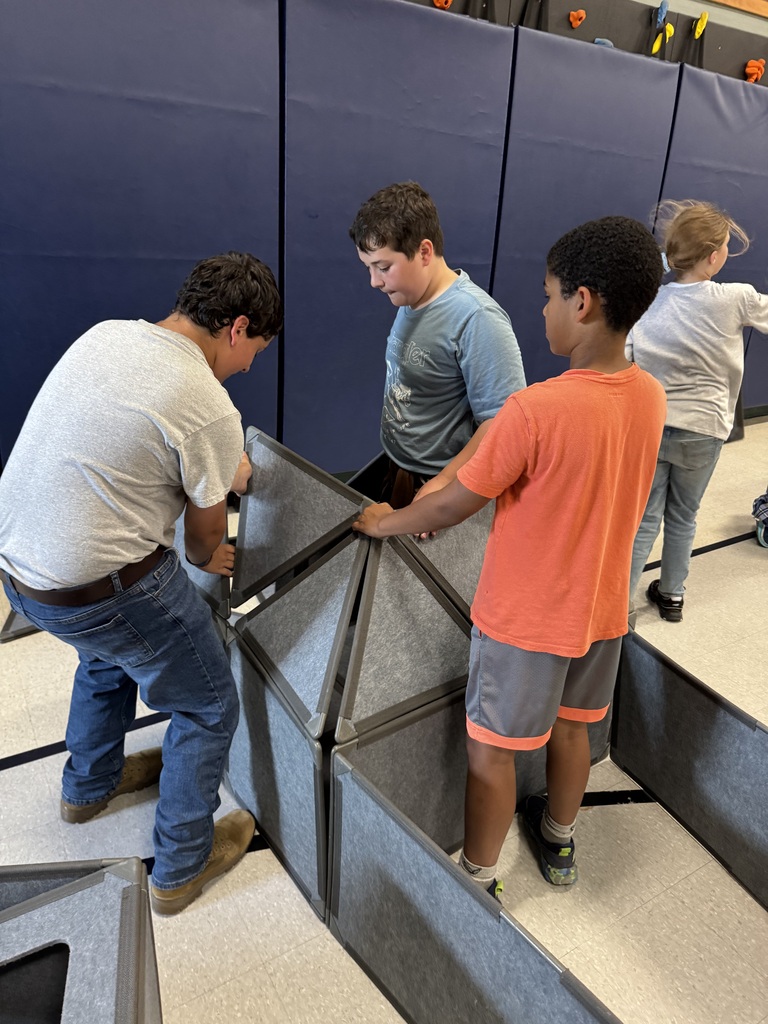Several children build with large gray modular panels in a gym or classroom space with blue wall mats in the background. Three children work together to attach triangular and rectangular gray panels into a tall structure while others move nearby.