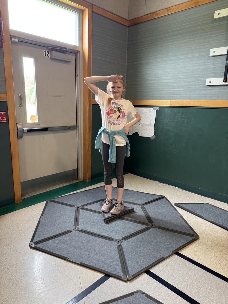 panels to form fort-like structures, while extra triangular and rectangular pieces lie scattered across the gym floor. IMG_6029.jpg A full-length shot of a young girl standing playfully on a flat geometric design constructed from the grey building panels on a gymnasium floor. The panels are arranged into a large, flat octagon with a small triangle in the center where she stands. She is wearing a white graphic t-shirt, grey leggings, and a light blue sweatshirt tied around her waist. She smiles at the camera, offering a playful military salute with her right hand. IMG_6021 (1).jpg A high-angle, close-up shot of a young boy peeking out from inside a small, triangular fort built from the grey modular panels. The structure is positioned against a dark green gym wall. The boy has dark hair, rosy cheeks, and a bright smile as he looks directly up at the camera from his cozy, makeshift hiding spot. IMG_6026.jpg A close-up shot looking through a square window cutout of a grey modular structure, framing three smiling boys crowded together inside. The boy on the left wears a grey shirt, the boy in the center foreground wears a black shirt and smiles widely with tilted head, and the boy on the right wears a bright orange t-shirt. The interior of the fort is dimly lit, creating a cozy clubhouse atmosphere.