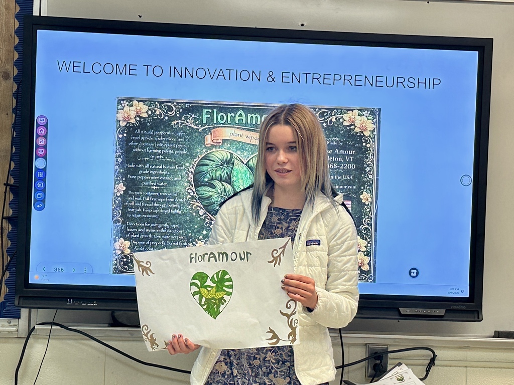 A student presents Flora Amour branding materials and product packaging concepts while standing in front of a classroom display showing the company’s promotional designs.