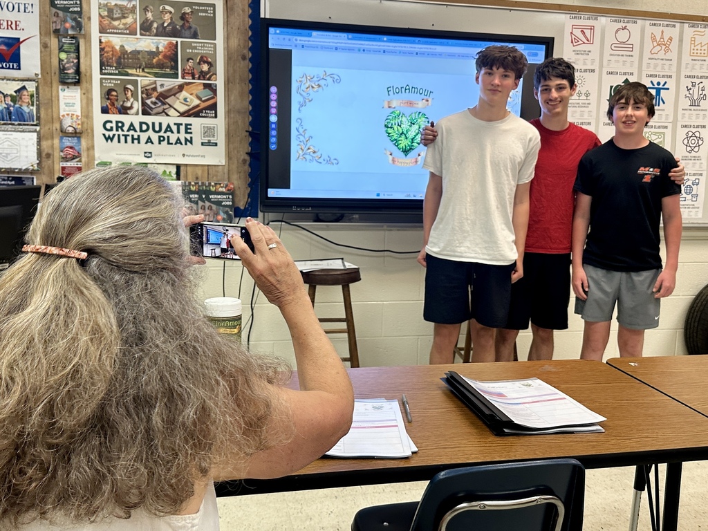 Sue Cook takes a photo of three students standing in front of a presentation screen displaying branding for Flora Amour plant wipes.