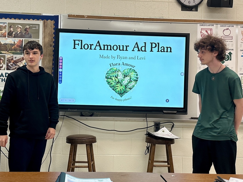 Two students stand in front of a presentation slide titled “FlorAmour Ad Plan,” explaining their marketing recommendations to classmates and a client.