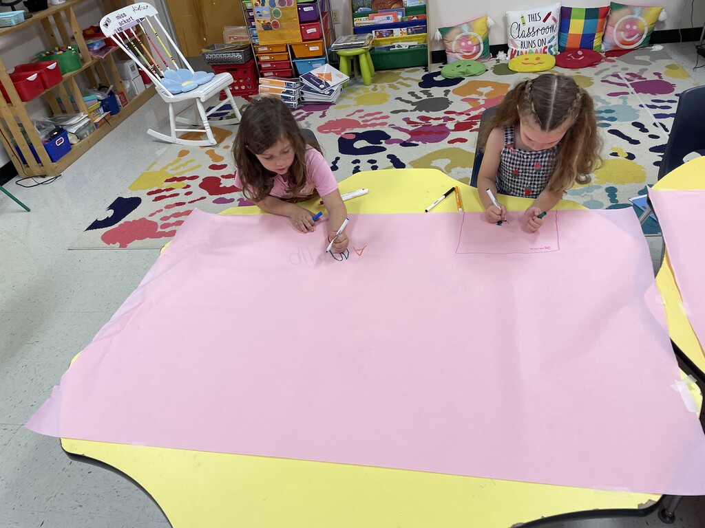 Two young girls sit side-by-side at a yellow table, working at the top edge of a large, mostly blank sheet of pink butcher paper. The girl on the left, wearing a pink shirt, is writing letters with a blue marker, while the girl on the right, with her hair in braided pigtails, carefully draws a small square box. In the background, a colorful handprint rug and a white rocking chair are visible.
