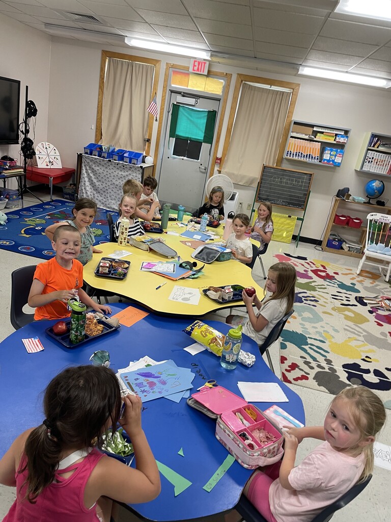 A bright, busy early childhood classroom where a group of about ten young children are sitting around interconnected blue and yellow kidney-shaped tables. Some children are eating lunch and snacks from trays and lunchboxes, while others are working on drawings and activity sheets with markers. The room is filled with colorful learning materials, including a handprint-patterned rug, bookshelves, and a whiteboard in the background. .