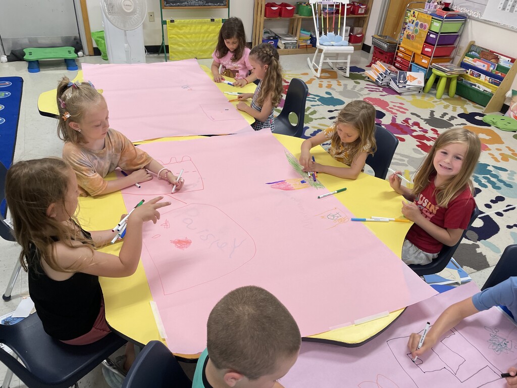 : A wide view of the classroom showing multiple groups of children engaged in a collaborative drawing project at yellow and blue tables. Each table is topped with a large sheet of pink paper. In the foreground, a group of girls use markers to draw and write their names on the paper, with one girl in a red shirt smiling directly at the camera.