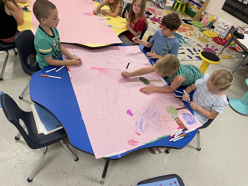 : A bright, busy early childhood classroom where a group of about ten young children are sitting around interconnected blue and yellow kidney-shaped tables. Some children are eating lunch and snacks from trays and lunchboxes, while others are working on drawings and activity sheets with markers. The room is filled with colorful learning materials, including a handprint-patterned rug, bookshelves, and a whiteboard in the background.