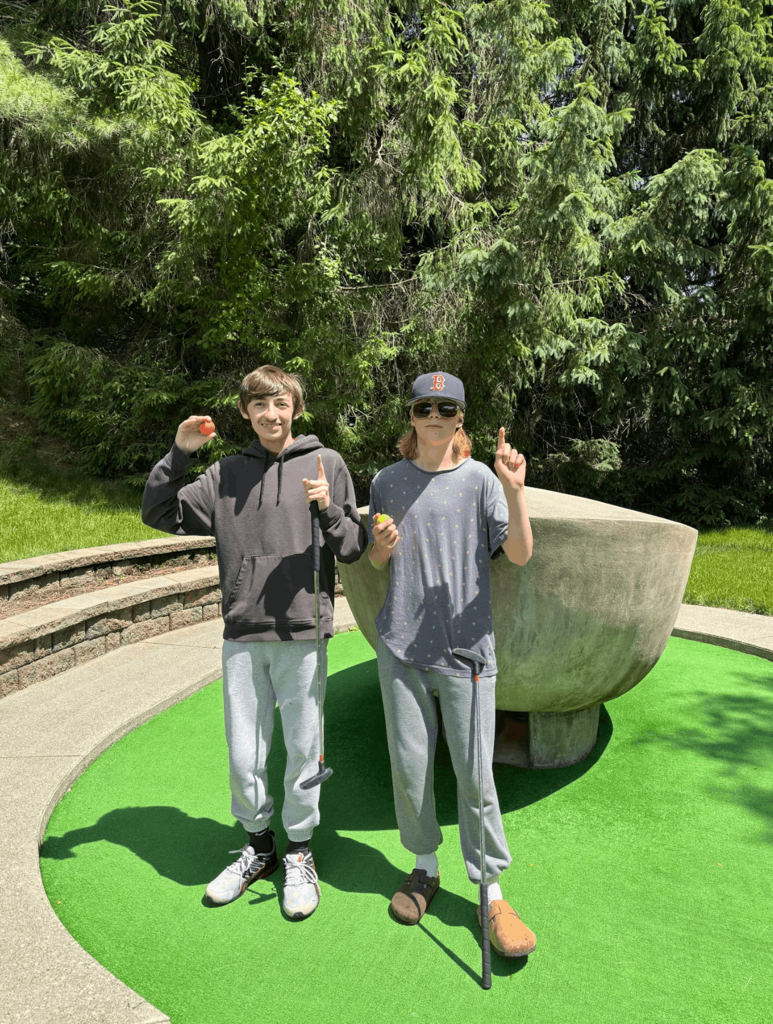 Two students celebrate a successful mini golf hole, holding golf balls and raising one finger while standing on a putting green.