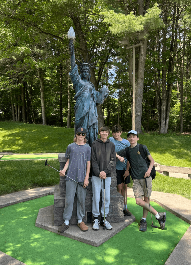 Four students pose with mini golf putters in front of a Statue of Liberty replica at a miniature golf course.