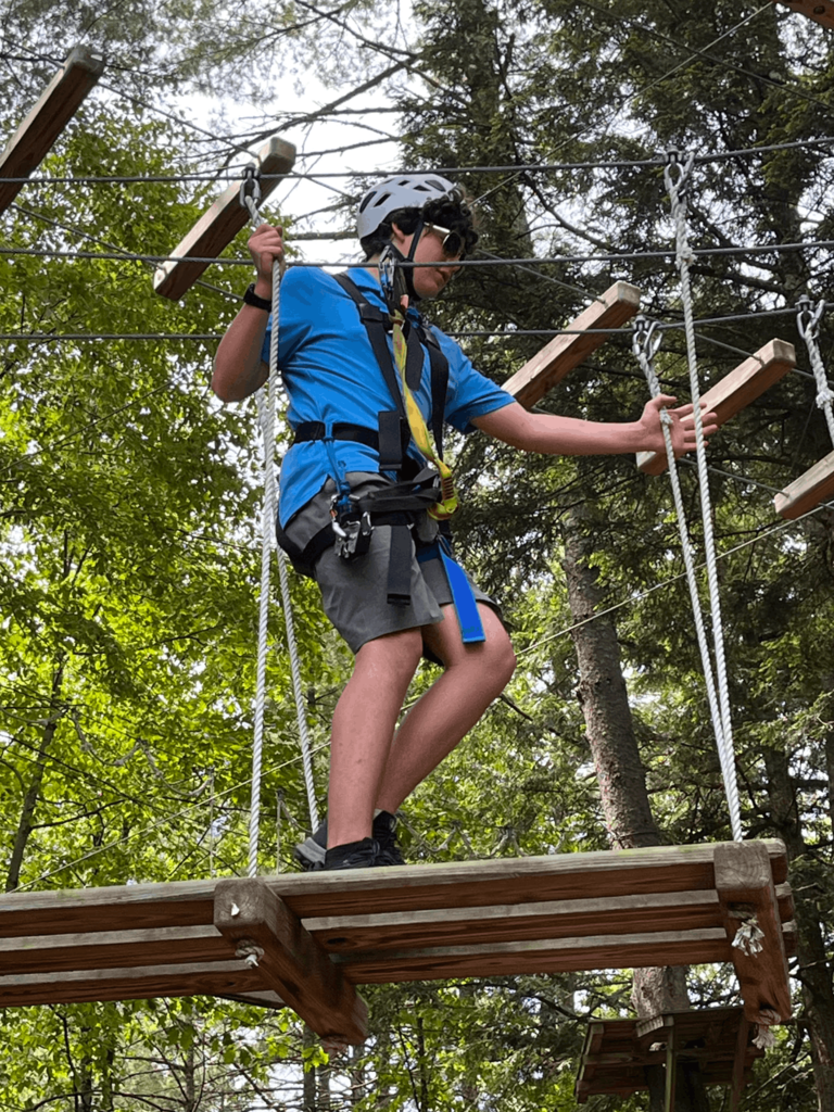 A student wearing a helmet and harness carefully balances across a suspended ropes course element high among the trees.