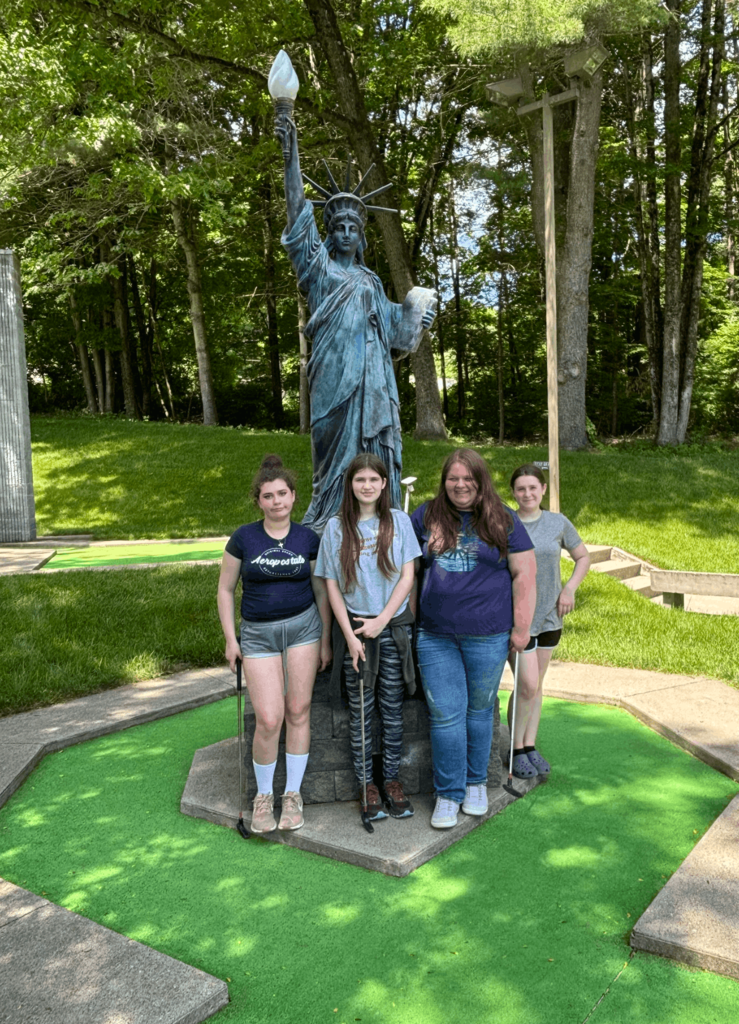 Four students stand smiling with mini golf putters in front of a Statue of Liberty replica on a miniature golf course.